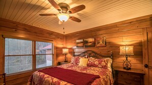 Une chambre dans une cabane en bois, avec un lit, deux tables de chevet, un ventilateur de plafond et une fenêtre avec des stores.