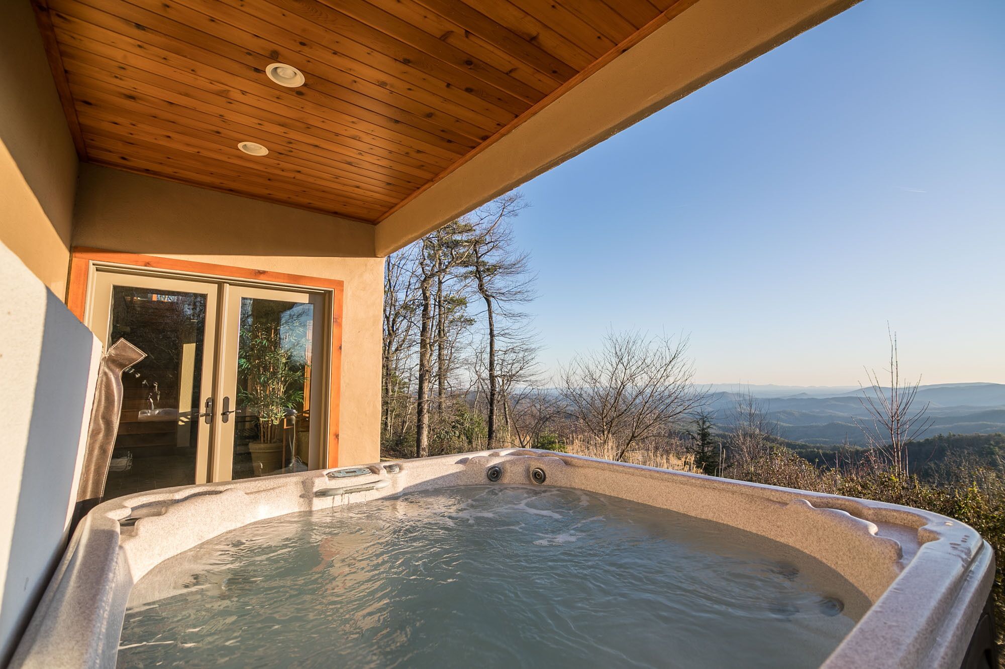 A hot tub on a balcony with a wooden ceiling, a glass door, and a view of trees and mountains.