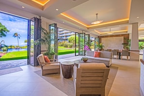 Un moderno vestíbulo de hotel con un amplio ventanal que da a una zona de piscina, una sala de estar con sofás y una mesa de centro, y un comedor con mesa y sillas.
