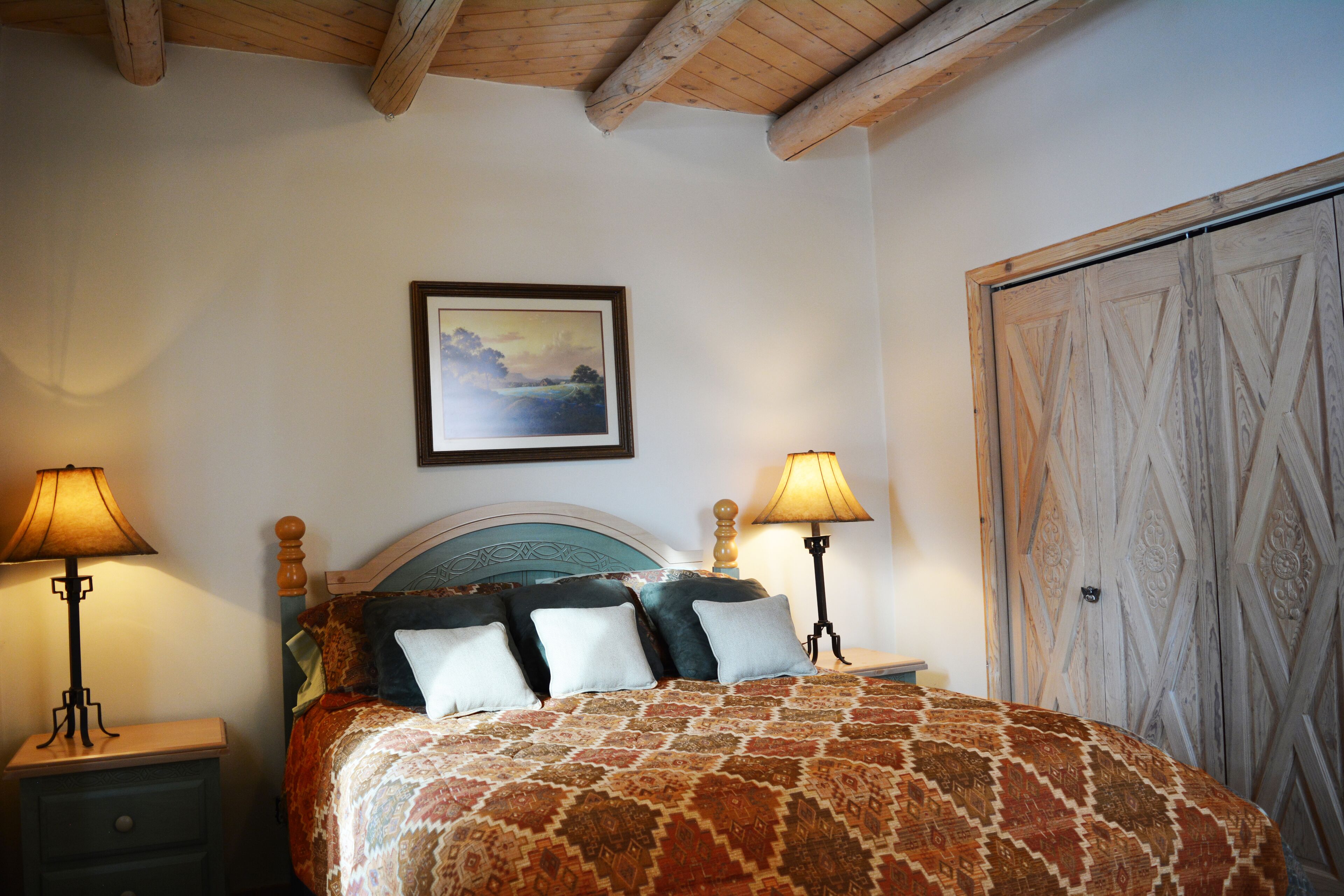 A bedroom with a bed, two bedside tables, a nightstand, a wooden wardrobe, and a framed picture on the wall.
