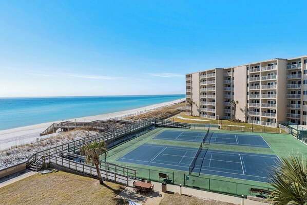 A tennis court adjacent to a beach, with a multi-story building in the background.