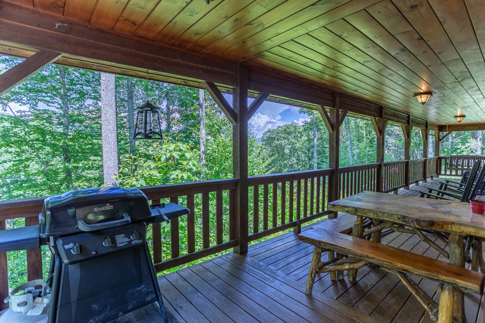 A covered outdoor area with a grill, picnic table, and benches, surrounded by a wooden deck and forest views.