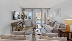 A modern living room with a sofa, coffee table, and TV. A balcony with a pool view is visible through the door. - 213- Unwind, Relax & Chill in this lovely Beachfront Condo w/ W&D,FREE Bch Sv (Destin)