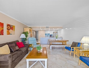 Un salon moderne avec un canapé, une table basse et des fauteuils. On aperçoit à l’arrière-plan un coin cuisine avec une table à manger et des chaises.