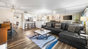 Un salon moderne avec un canapé en cuir noir, une table basse en bois et un coin cuisine à l’arrière-plan.
