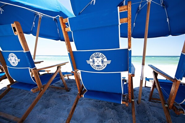 Strandstoelen met blauwe zonnedoeken en houten frames, op een zandstrand met de oceaan op de achtergrond.