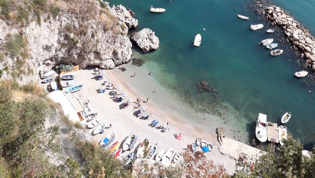 Uma área costeira com barcos atracados perto da praia, uma faixa de areia e falésias rochosas.
