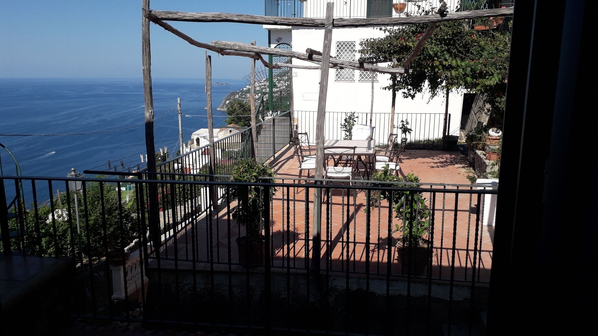 A balcony with a view of the sea, a white building, and a fenced patio area with outdoor furniture.