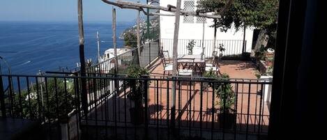 A balcony with a view of the sea, a white building, and a fenced patio area with outdoor furniture.