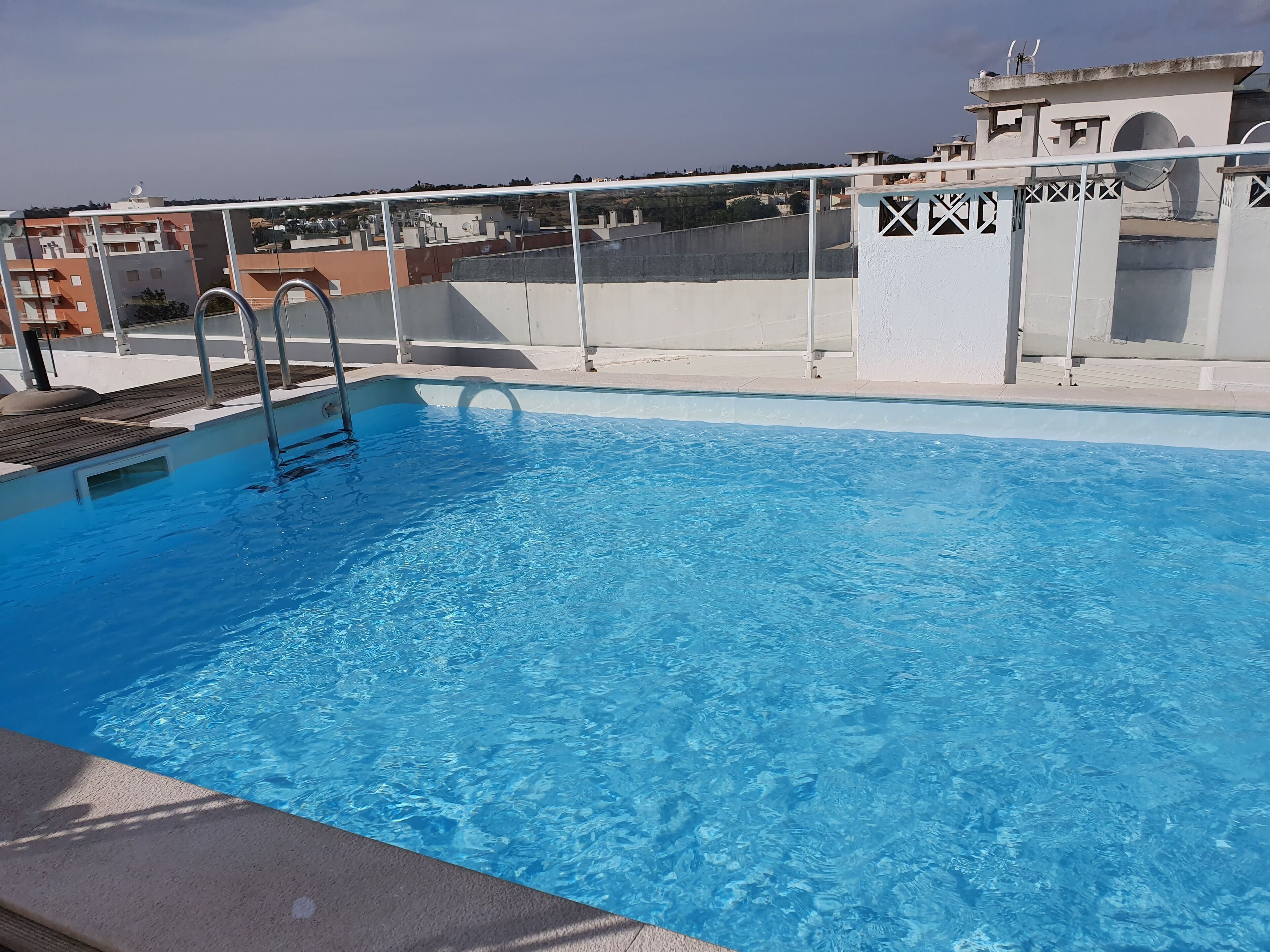 A rooftop swimming pool with a metal ladder, surrounded by buildings and a clear sky.