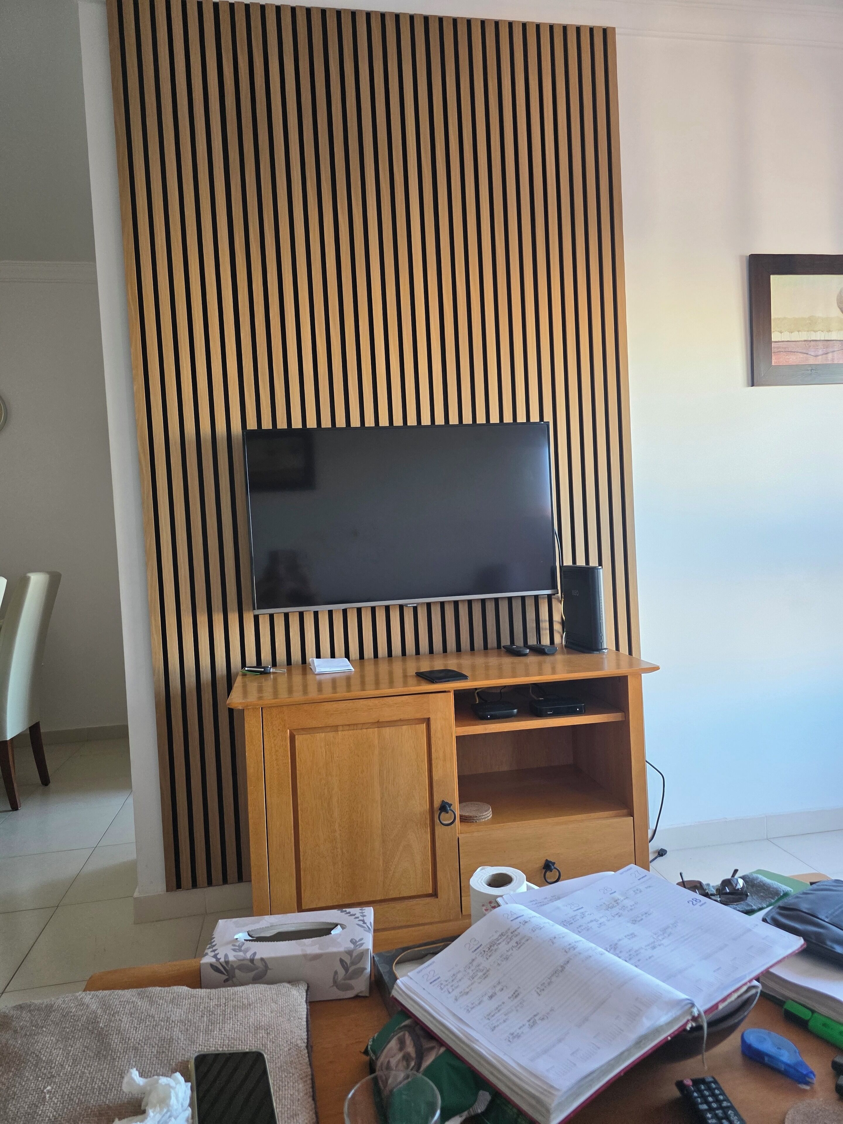 Une pièce comprenant un meuble de rangement en bois abritant un téléviseur à écran plat, une console de jeux et un haut-parleur. On y trouve également un bureau avec un ordinateur portable, un carnet et divers fournitures de bureau.
