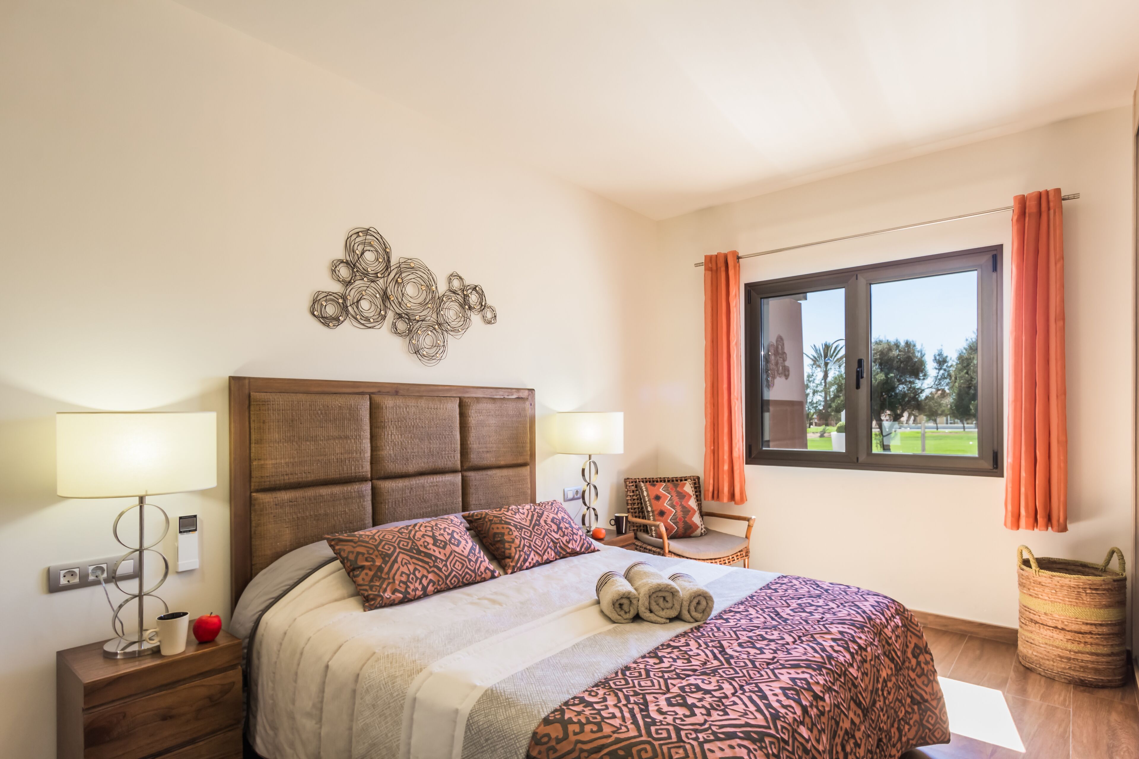 A neatly made bed with patterned bedding, a bedside table with a lamp, a decorative wall piece, and a window with curtains.