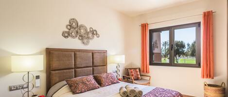 A neatly made bed with patterned bedding, a bedside table with a lamp, a decorative wall piece, and a window with curtains.