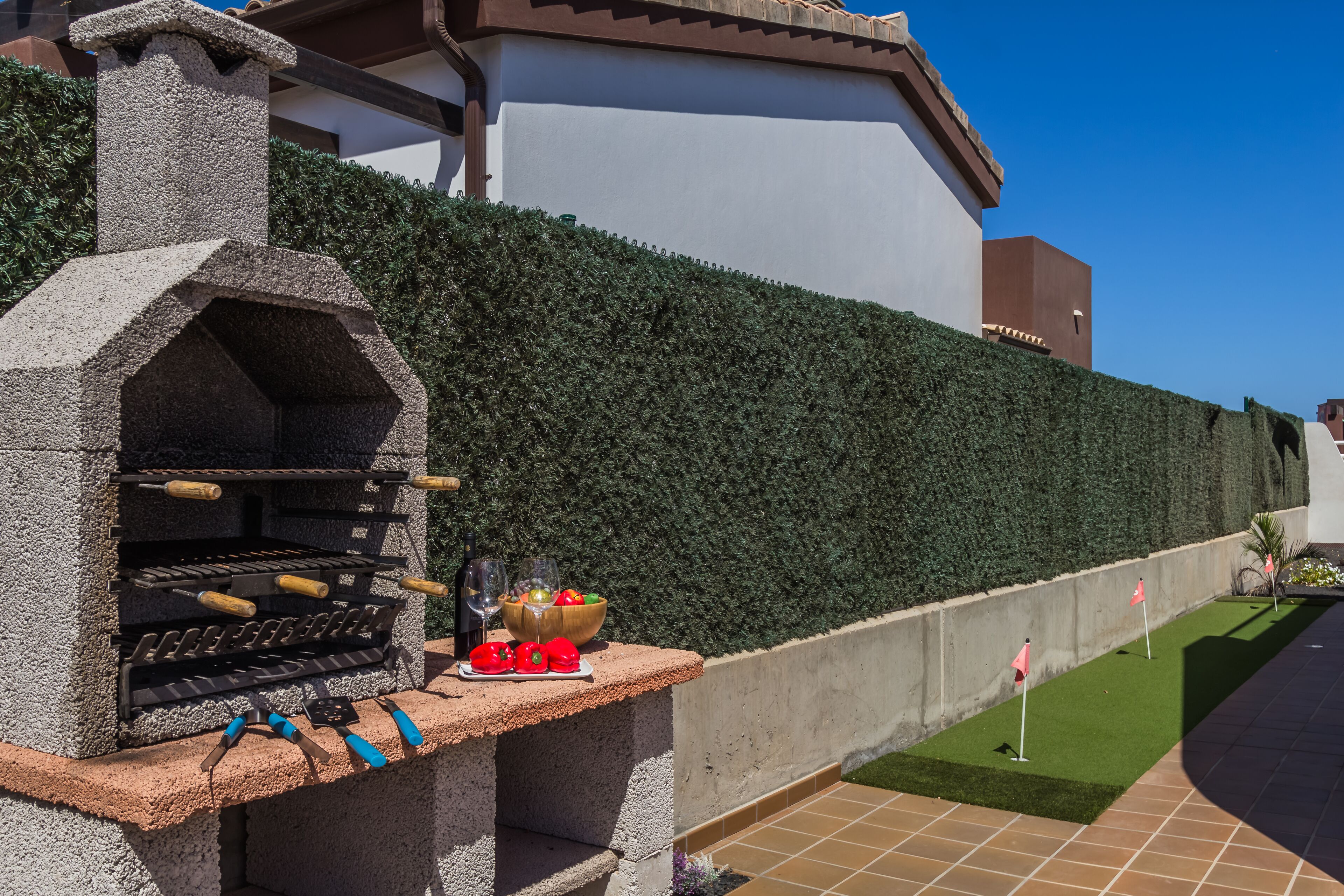 Een stenen barbecue met tuingereedschap en een schaal fruit, voor een haag en een gebouw.