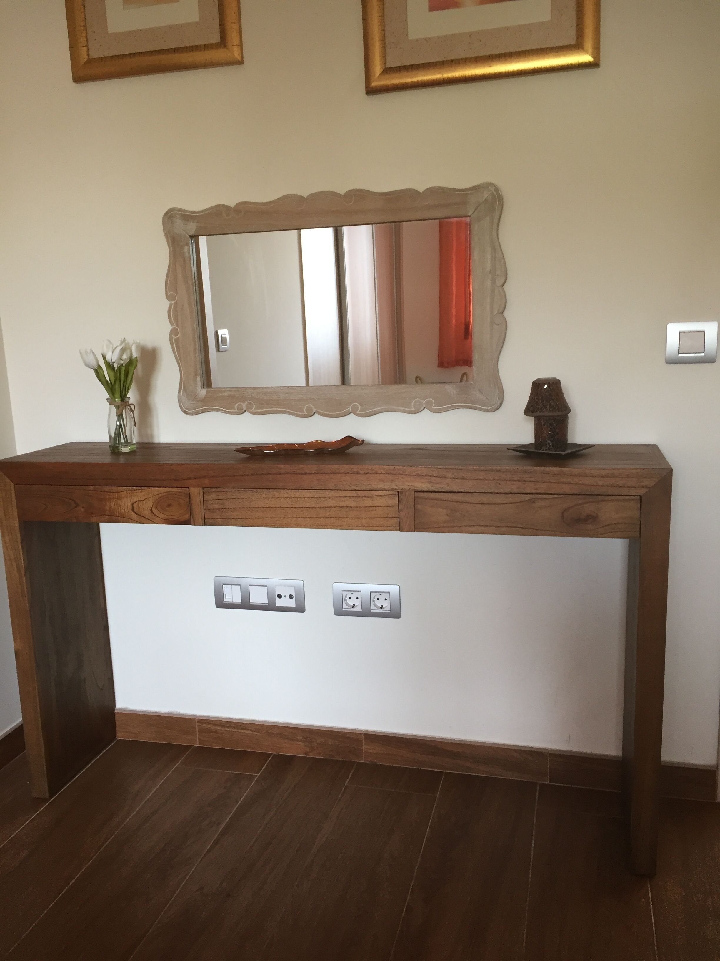 A wooden vanity with a mirror, a vase of flowers, and a lamp.