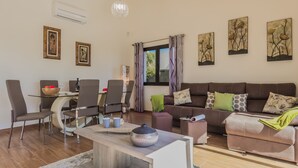 A modern living room with a glass dining table, brown leather chairs, a brown sofa with patterned cushions, and artwork on the walls.