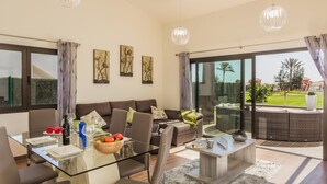 A modern living room with a glass dining table, brown sofa, and large windows.