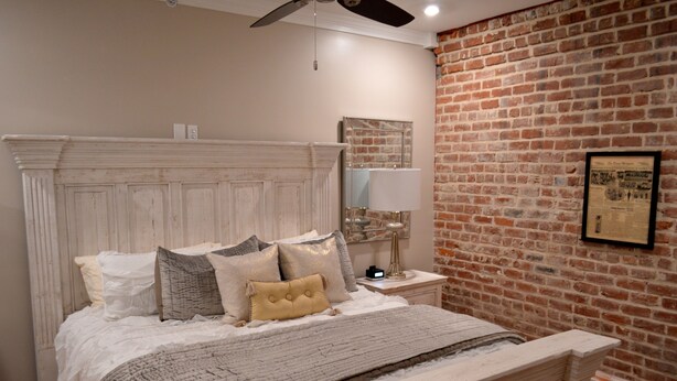 A bedroom with a brick wall, a large bed with multiple pillows, a nightstand with a lamp, and a ceiling fan. - Local Culture themed Historic luxury condo, 2 blocks from French Quarter (New Orleans)