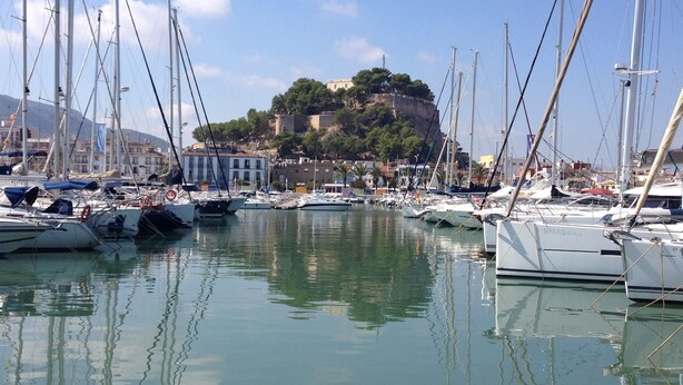 A marina with multiple sailboats docked, a historic building on a hill, and calm water reflecting the boats.