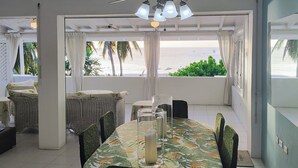 A dining area with a table set for four, surrounded by wicker chairs, a ceiling fan, and a view of the beach.