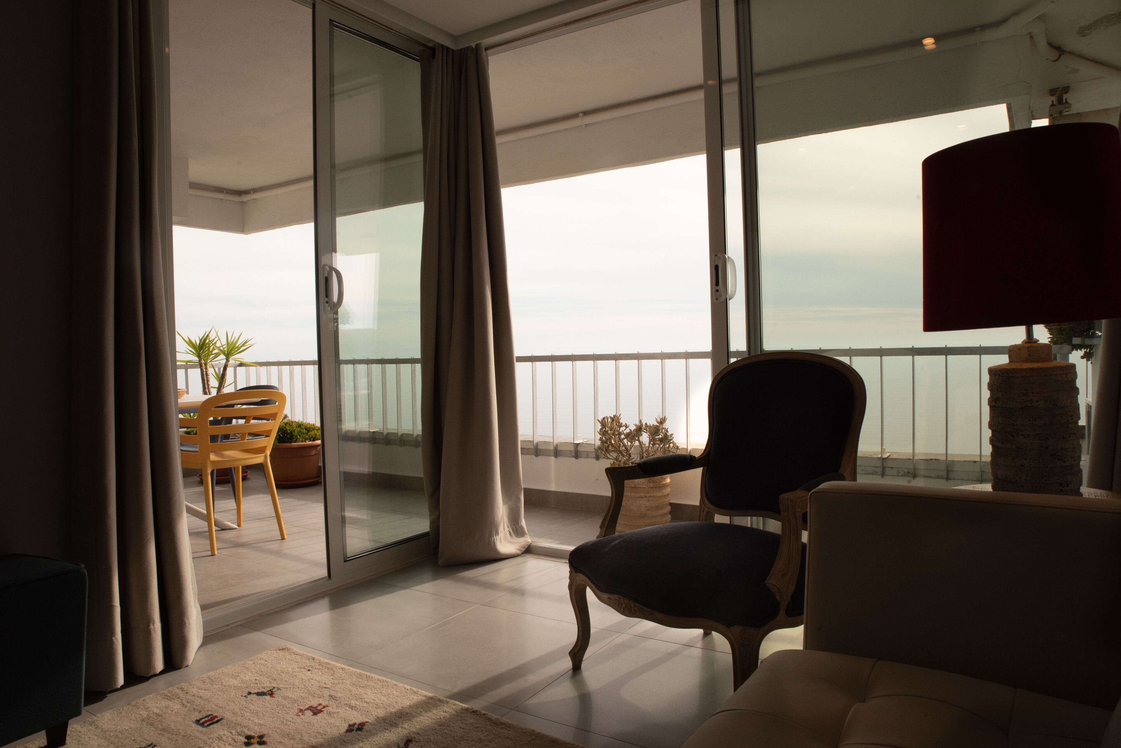 A balcony with a view of the sea, a chair, a table, and a lamp.