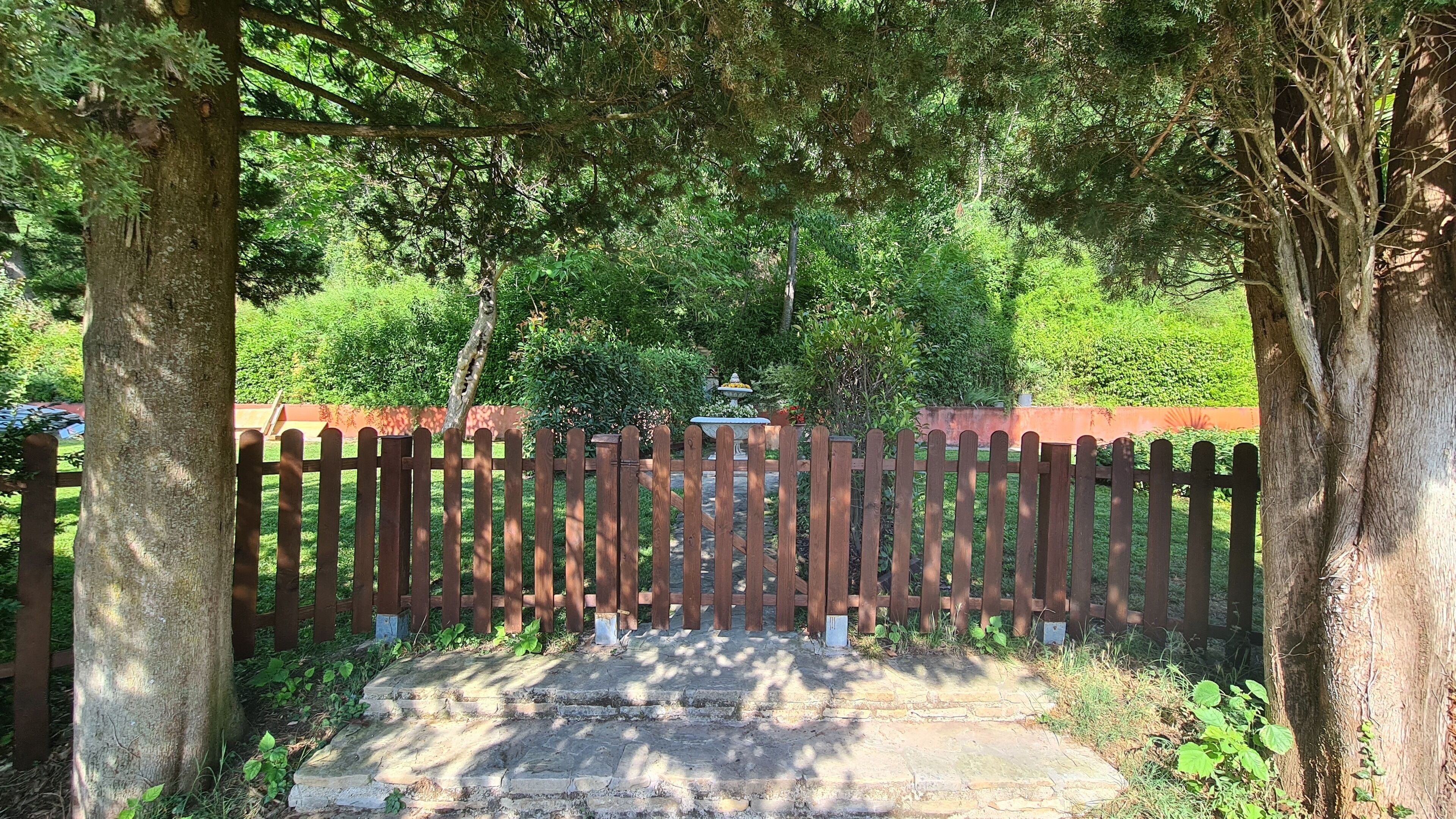 Une porte en bois mène à un sentier pavé de pierres, bordé d’une végétation luxuriante et d’arbres.