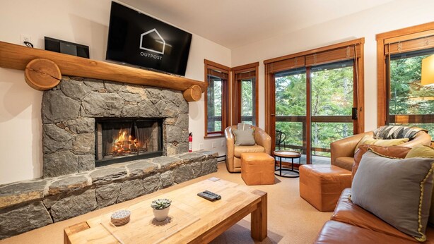 A cozy living room with a stone fireplace, leather furniture, and large windows offering a view of the outdoors. - Ski in/out | BBQ | Complex pool & Hot tub | Garage (Whistler)