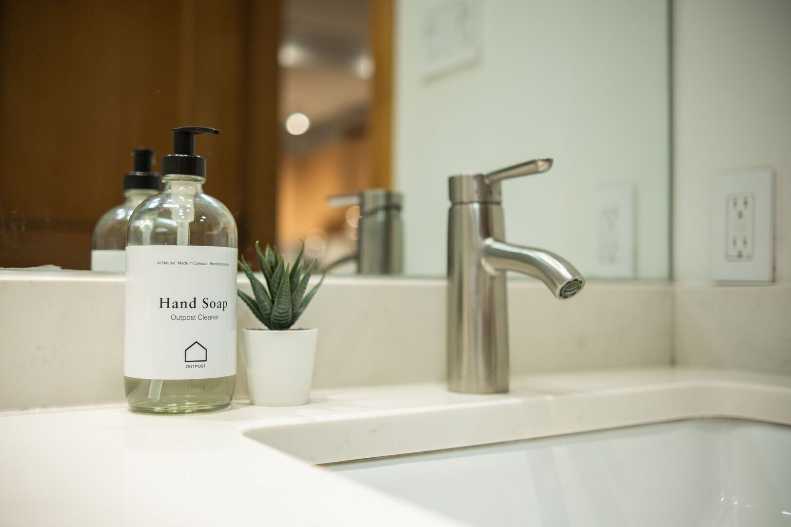 A bathroom sink with a hand soap bottle labeled "Outpost Cleaner" and a small potted plant.