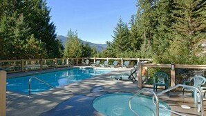 A pool area with lounge chairs and a wooden deck, surrounded by trees and mountains in the background. - Ski in/out | BBQ | Complex pool & Hot tub | Garage (Whistler)