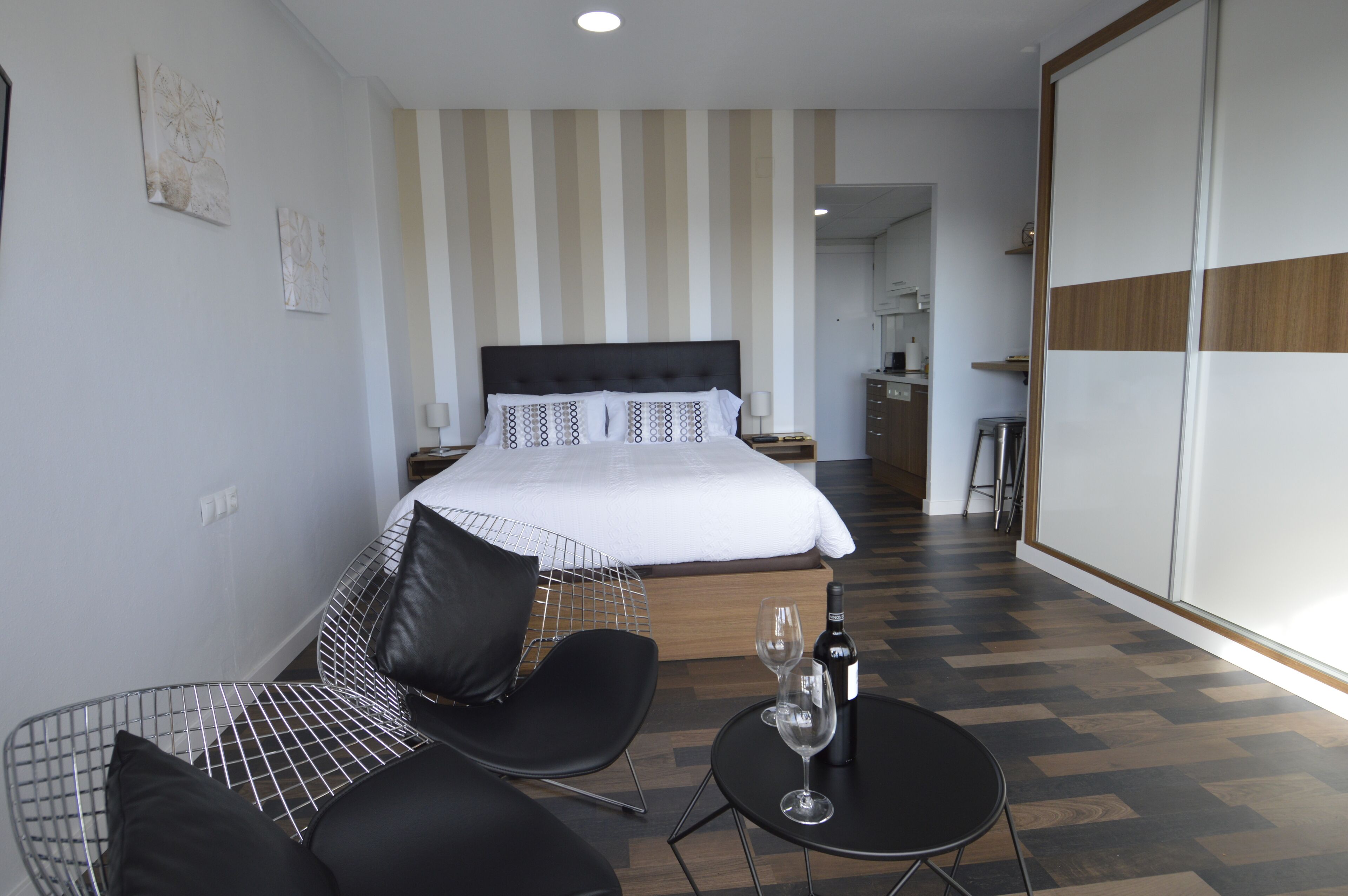 Habitación de hotel con cama, una silla, una mesita con una copa de vino y vista a la cocina.