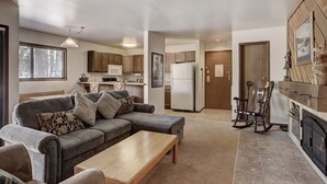 A cozy living room with a sofa, coffee table, and a fireplace. A kitchen with wooden cabinets and a refrigerator is visible in the background. - Ground Floor Condo, Courtyard View, Pool & Hot Tub, Shuttle to Slopes (Keystone)