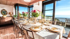 A dining area with a long table set for a meal, large windows with a view of the ocean, and wooden ceiling beams.
