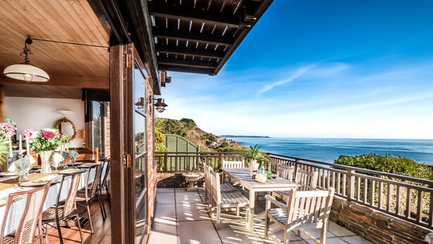 A balcony with a wooden table and chairs, a view of the ocean, and a stone wall.