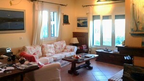 Un salon avec un canapé, une table basse et une cheminée. Il y a des fenêtres avec des rideaux, un tableau au mur et une lampe sur une table d’appoint.