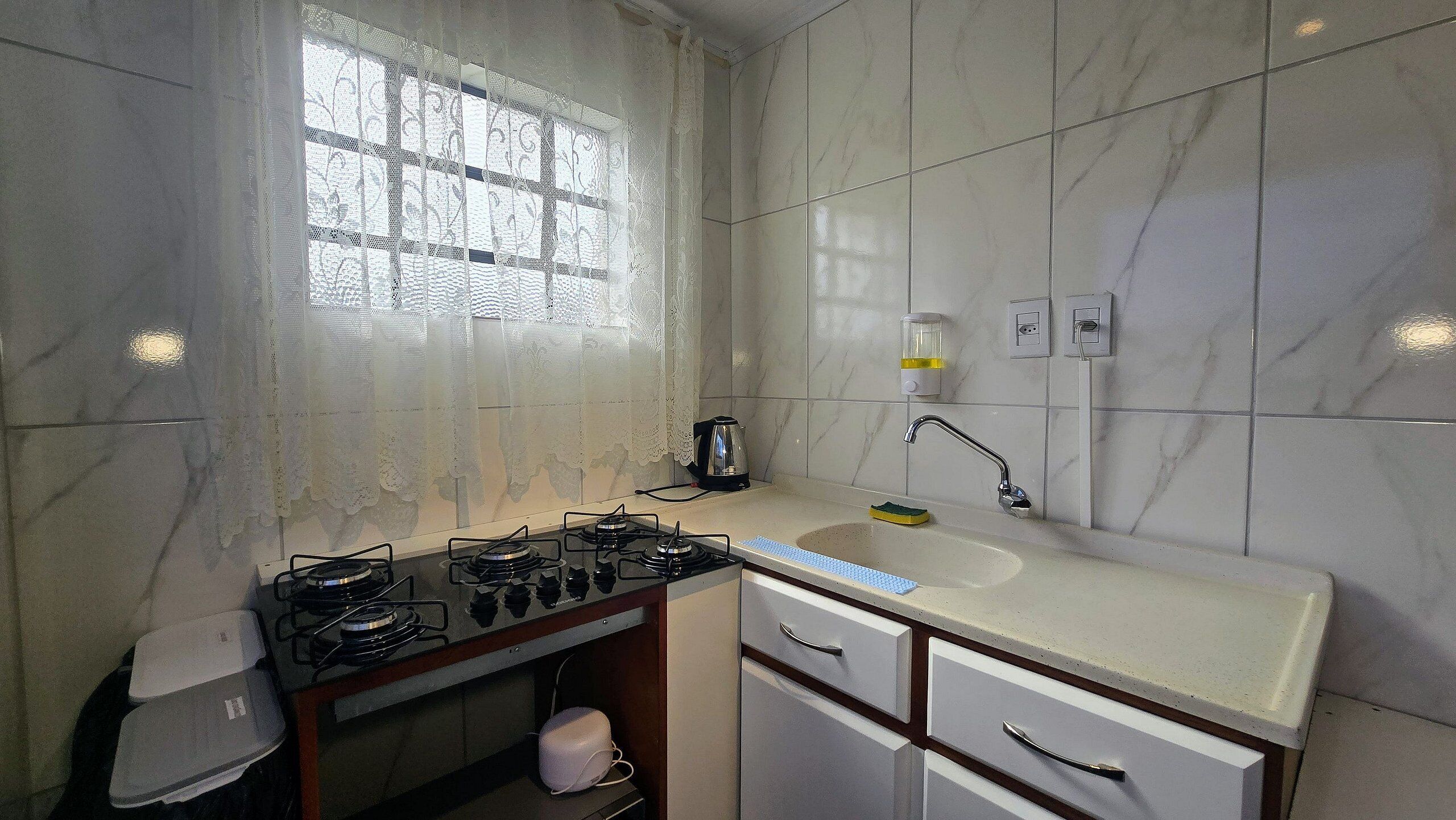 A kitchen with a gas stove, a sink, a window with lace curtains, and a coffee maker.