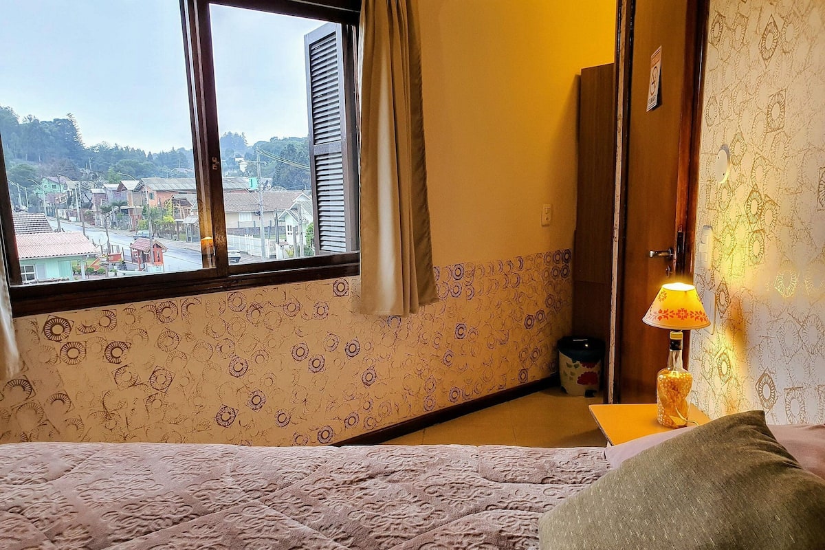 A hotel room with a bed, a window with a view of a town, a lamp, and patterned wallpaper.