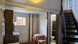 Une petite salle de bain comprenant des toilettes, un lavabo et une petite table avec une chaise. Un tabouret en bois est placé près de la table.