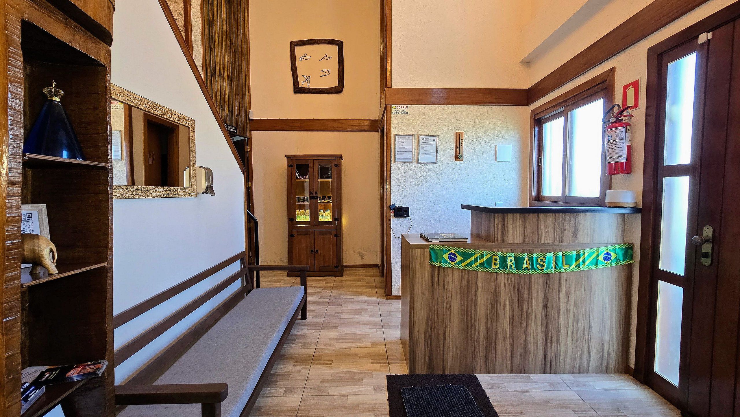 A reception desk with a Brazilian flag, a wooden bench, a door with glass panels, and a wall clock.
