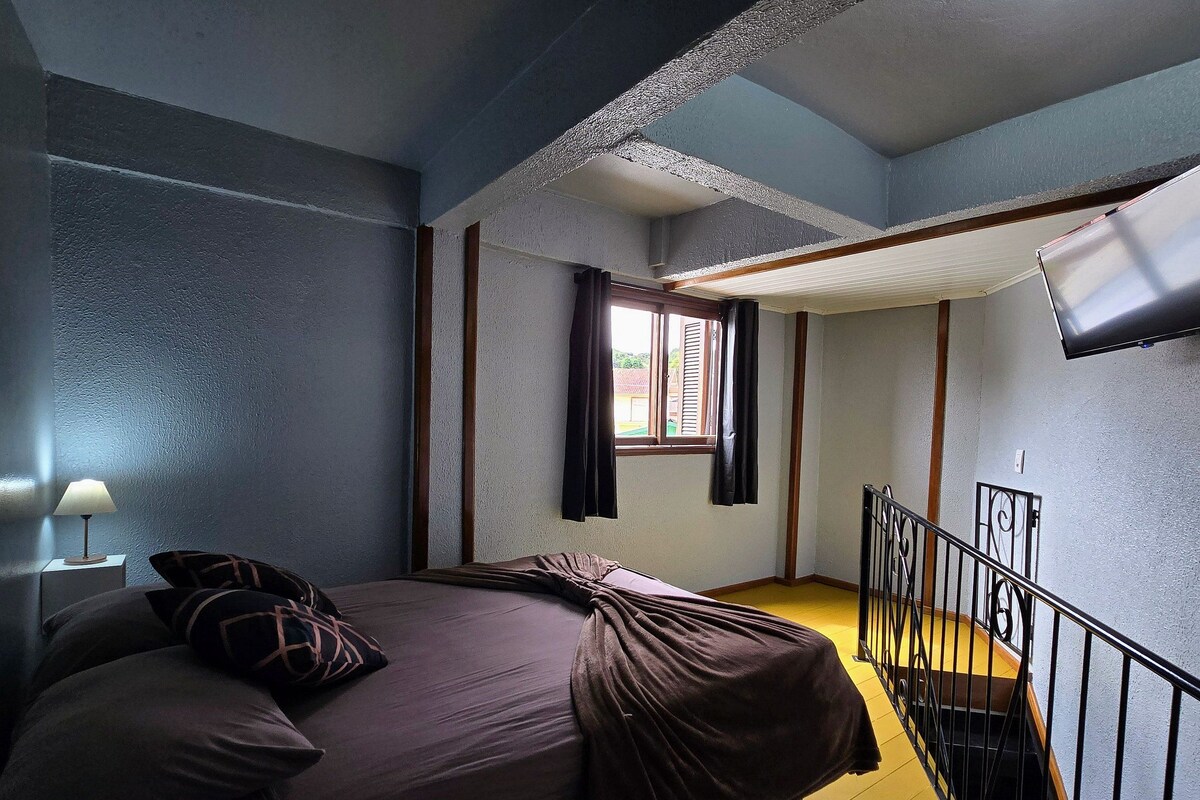 A bedroom with a bed, a nightstand, a window with shutters, a staircase, and a TV mounted on the wall.