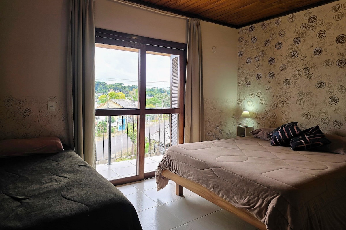 A bedroom with a large window, a bed, a bedside table with a lamp, and a view of the outside.
