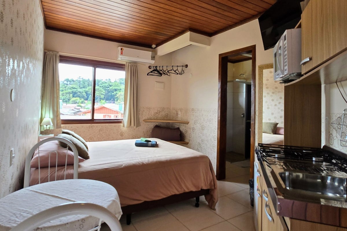 A compact room with a bed, a small table, a kitchenette, and a window with curtains.