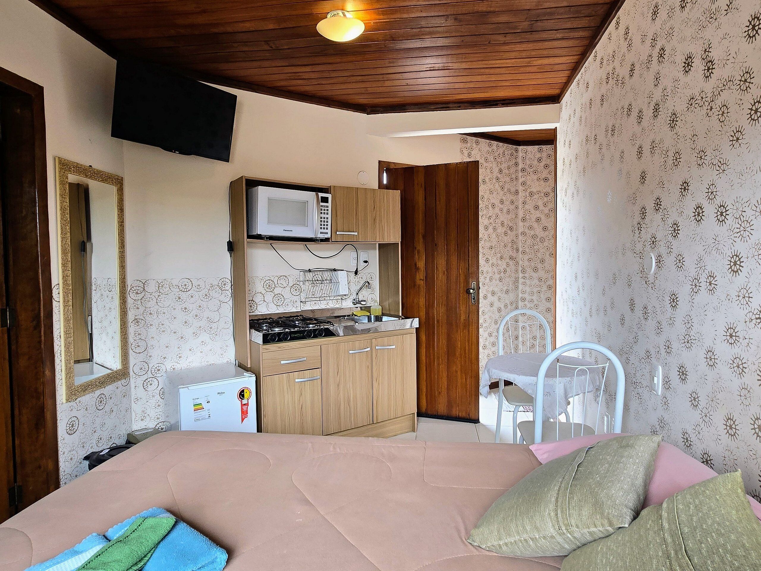 A compact bedroom with a bed, a small kitchenette, a TV mounted on the wall, and a mirror.