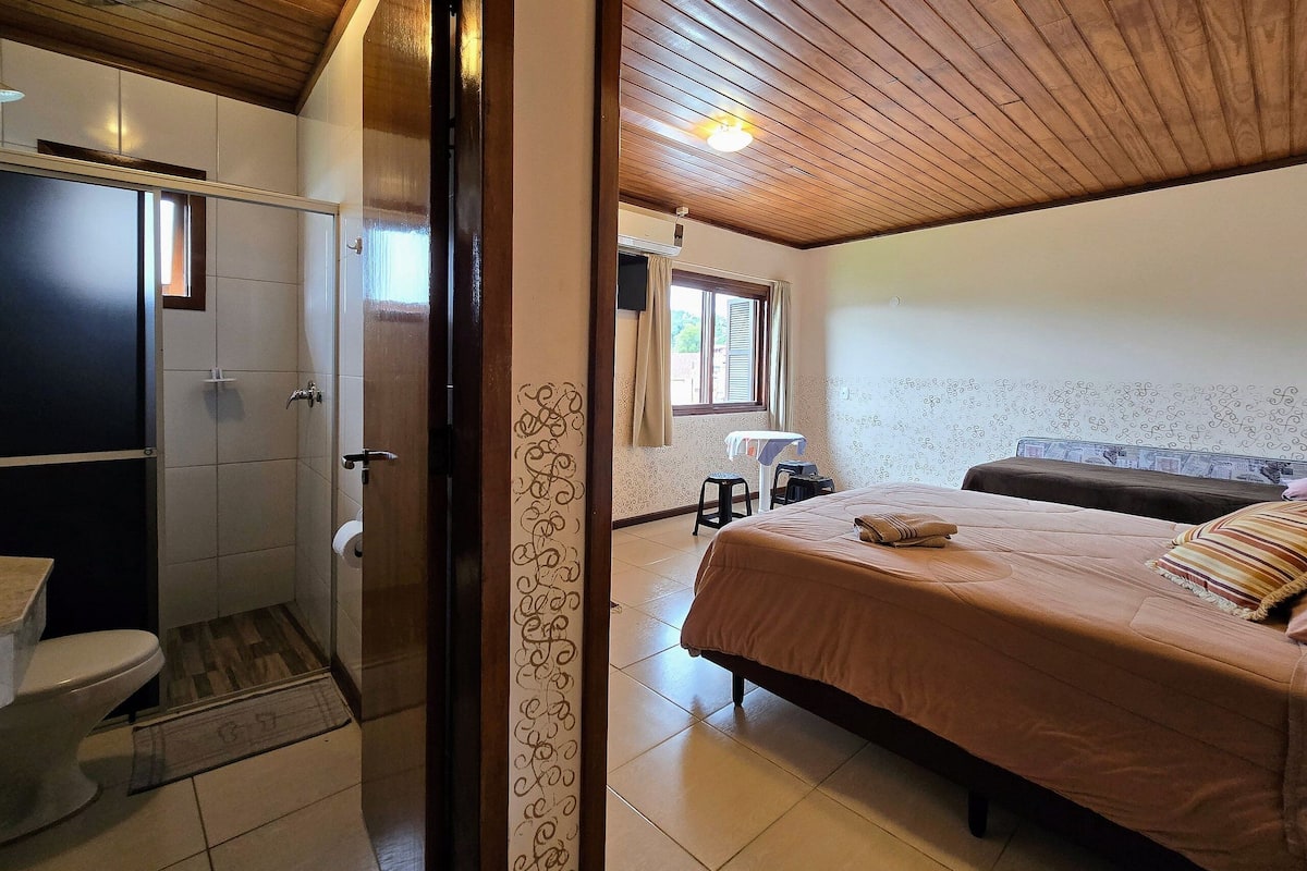 A bedroom with a wooden ceiling, a bed with a brown bedspread, a window with curtains, and a small table with a chair.