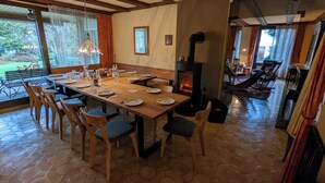 Zona pranzo con un lungo tavolo di legno apparecchiato per sei persone, circondato da sedie. Si vede una stufa a legna nell'angolo e la stanza ha ampie finestre con tende.