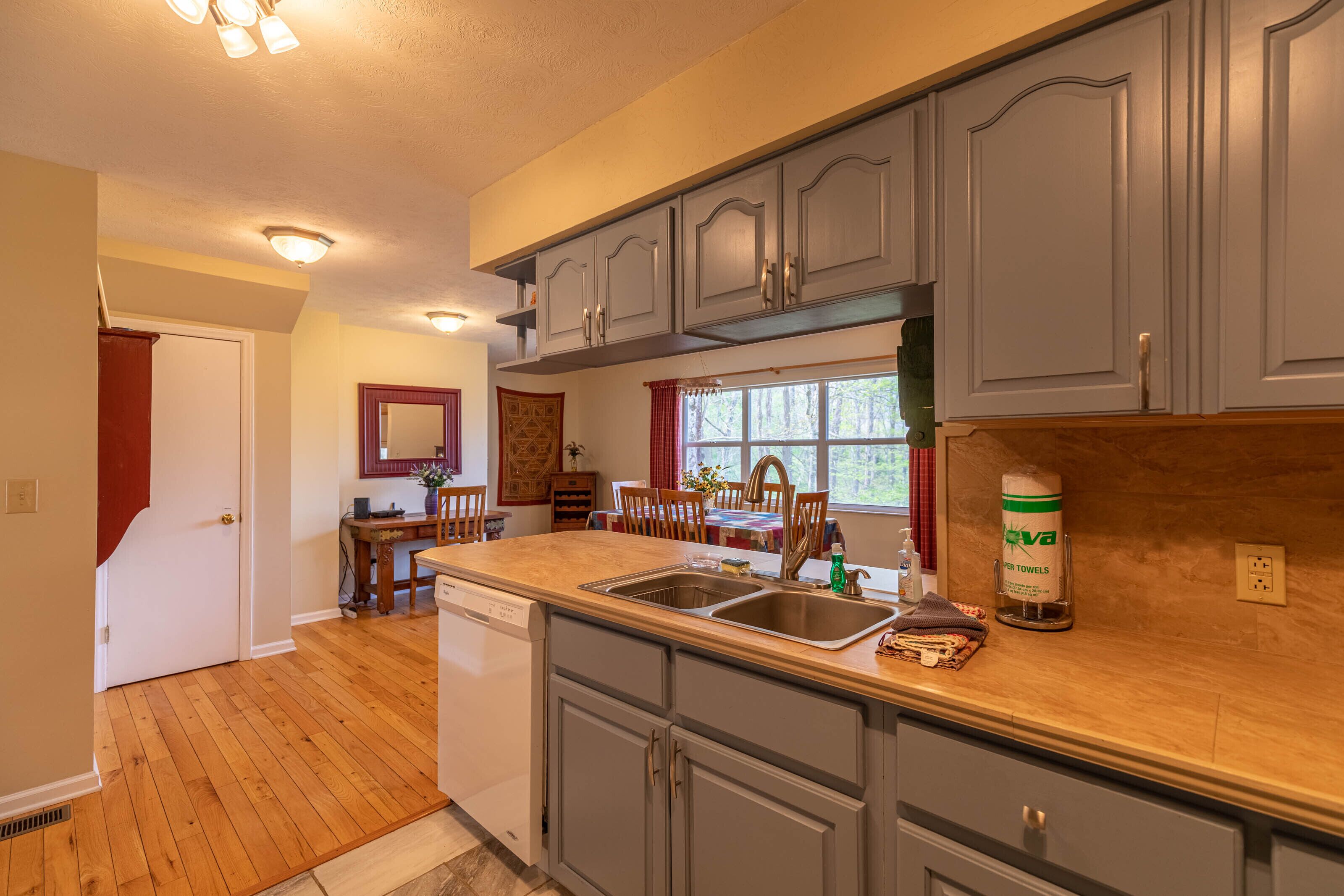 A kitchen with wooden flooring, grey cabinets, a dishwasher, and a window with red curtains.