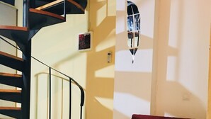 Una sala de estar con un sofá rojo, una mesa de centro de cristal con manzanas, una escalera en espiral y una lámpara fijada a la pared.