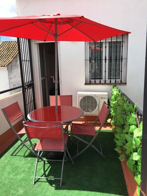A small outdoor patio with a round table and red chairs, a red umbrella, and a green plant.