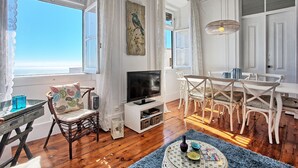 A spacious living room with a large window overlooking the sea, a dining area with white furniture, and a television on a white stand. - RM3 - River View | Alfama | AC | 4BR | 3 BATH Sleeps 9 (Lisbon)