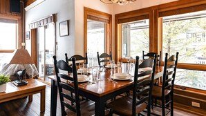 A dining area with a wooden table set for six, surrounded by chairs, with a chandelier above and a view of trees outside.