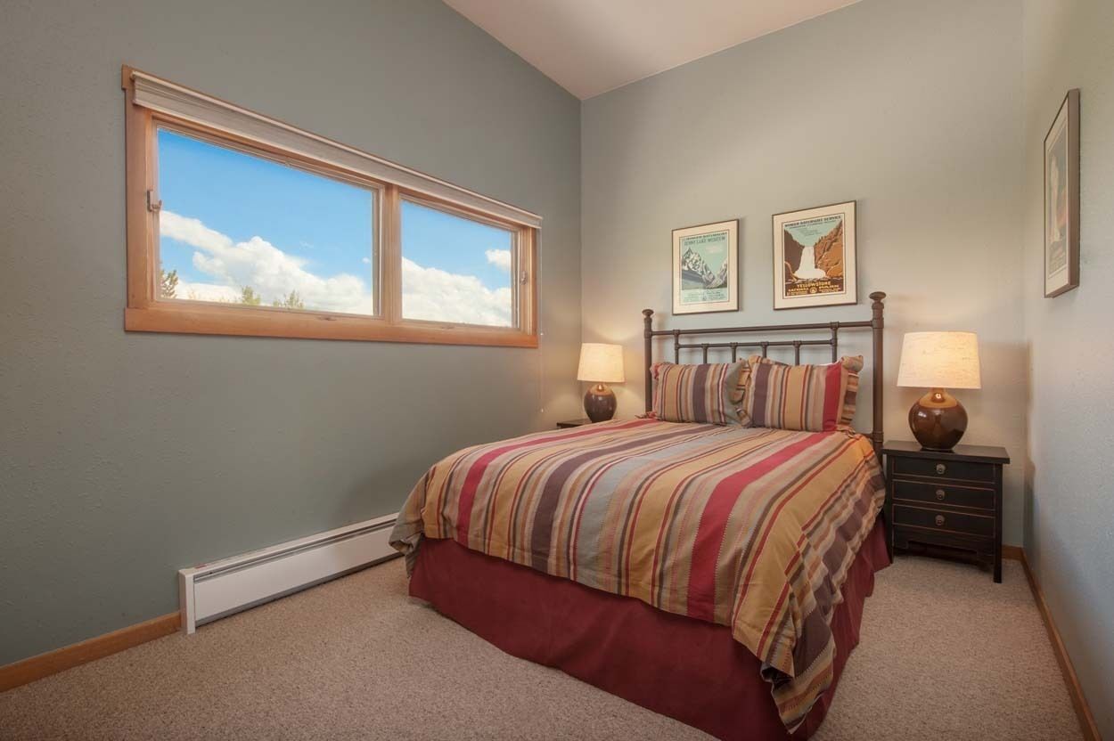 Un dormitorio con un ventanal grande, una cama con ropa de cama a rayas, dos mesitas de noche con lámparas y dos cuadros en la pared.